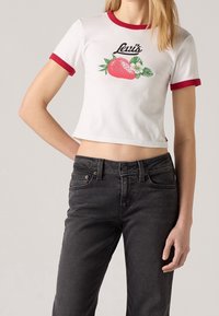 Camiseta blanca recortada con acentos rojos, que presenta un gráfico de una fresa y el logo de "Levi's" en negro. Llevada con jeans oscuros.