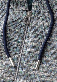 Veste zippée en tissu texturé avec un motif bleu et blanc, éléments en métal argenté, et cordons de serrage contrastants en marine.