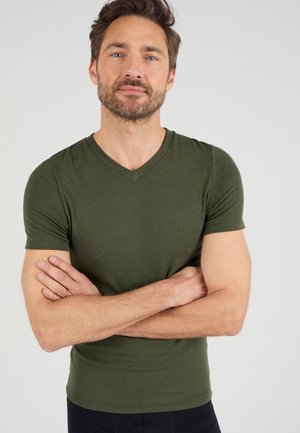 T-shirt vert olive à manches courtes et col en V, fabriqué dans un matériau doux, au design ajusté et à la texture lisse. La personne a les bras croisés dans une pose neutre.