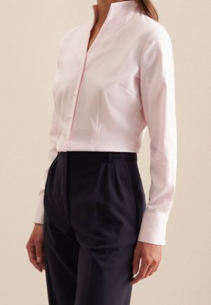 Femme portant un haut court rose pâle à manches longues et un pantalon noir plissé taille haute, sur un fond neutre.