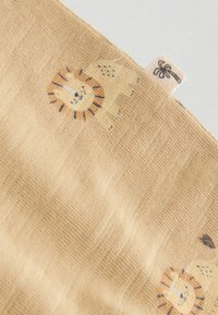 Svijetlosmeđa tkanina s uzorkom malog lavljeg lica i tkanom etiketom na kojoj se nalazi crna ikona palme.