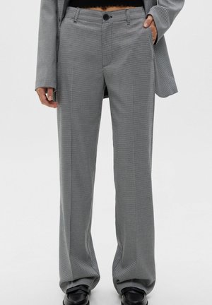 Person iført små sort-hvide ternede bukser, sorte loafers og en matchende ternet blazer med den ene hånd i lommen.