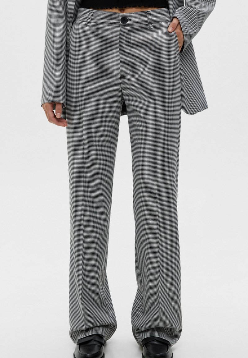 Personne portant un pantalon à petits carreaux noirs et blancs, des mocassins noirs et un blazer à carreaux assorti, une main dans la poche.