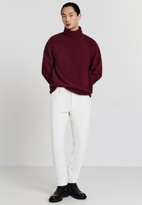 Burgunderfarbener gestrickter Rollkragenpullover, cremefarbene Hose und schwarze Stiefeletten. Der Pullover hat einen gerippten Kragen; die Hose hat eine subtile Textur.