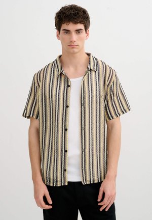 Joven con camisa de rayas de manga corta abotonada sobre una camiseta blanca sin mangas, de pie contra un fondo liso y claro.