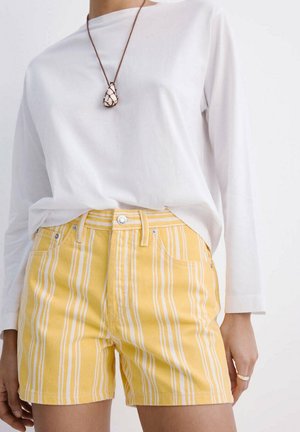 Mujer con camisa blanca de manga larga, pantalones cortos a rayas verticales amarillas y blancas, collar con colgante de concha y anillo en la mano derecha.