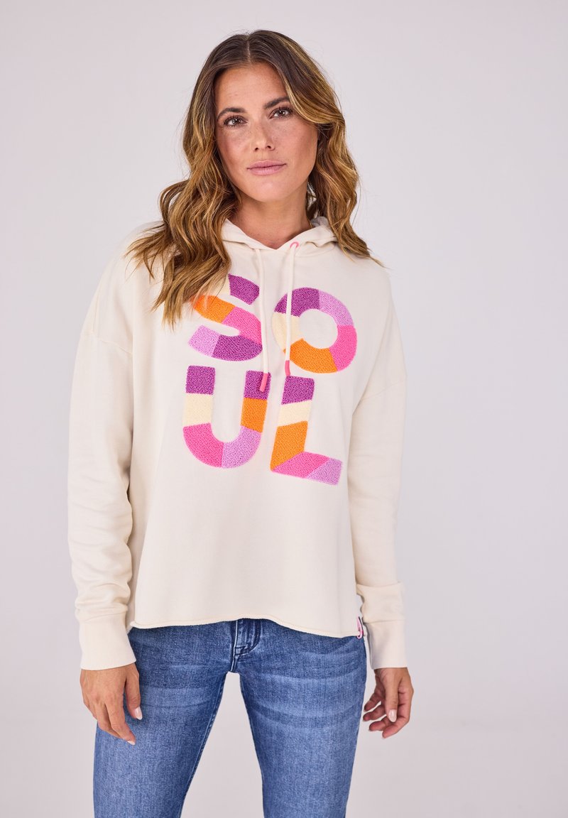 Cremfarbener Hoodie mit einem großen, mehrfarbigen "SOUL"-Grafik auf der Vorderseite. Er verfügt über eine Kapuze mit Kordelzug und eine weiche Textur. Kombiniert mit blauen Jeans.