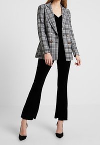 Dvojité sako v černé, šedé a bílé kostce se zlatými knoflíky, spárováno s černým sametovým overálkem a černými vysokými podpatky.