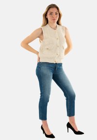 Chaleco sin mangas de punto en color crema con textura acanalada, presenta dos bolsillos frontales y botones dorados, combinado con jeans ajustados azules y tacones negros.