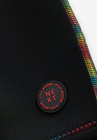 Black fabric with multicolor overlock stitching and a round black patch with red text "NEXT" and "STAY ACTIVE" around the edge.