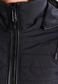 Chaqueta acolchada oscura con cuello alto, cremallera negra y textura de tela sintética, con detalles de cuero suave en la cremallera y los bordes.
