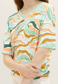 Femme portant un t-shirt à manches courtes et col en V avec un motif abstrait ondulé vert, orange, moutarde et bleu, les bras croisés à la taille.