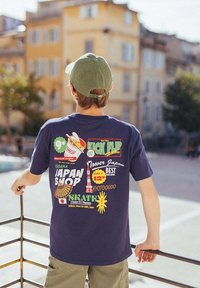 Garçon portant une casquette verte et un t-shirt graphique bleu marine avec un motif sur le thème du Japon, debout à l'extérieur tenant une rambarde en métal avec des bâtiments en arrière-plan.