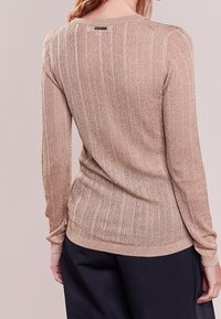 Suéter de manga larga acanalado en color dorado claro, con forma ajustada y textura suave. Etiqueta visible en la parte posterior del escote, combinado con pantalones oscuros.