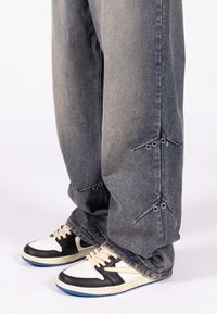 Φαρδιά τζιν με ξεθωριασμένη μπλε επίστρωση, διακοσμημένα με τρουκς και μοναδικές τριγωνικές κοπές κοντά στο τελείωμα, συνδυασμένα με μαύρα και άσπρα αθλητικά παπούτσια.