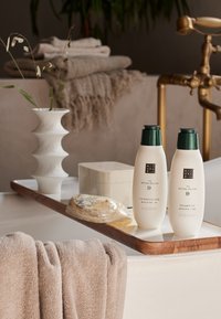 Weiße Shampoo- und Spülungsflaschen mit grünen Kappen auf einem Holztablett. Auch eine durchsichtige Seifenstücke, eine weiße Keramivase und ein beiger Handtuch sind enthalten.