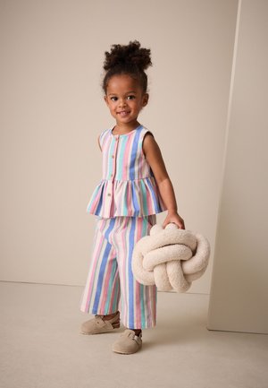 Ung pige smiler, iført ærmeløs pastelstripet outfit, holder en beige knyttet plyspude, står mod en neutral baggrund.