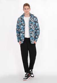 Jachetă ușoară cu un model de frunze de palmier în nuanțe de albastru, negru și alb; design ajustat cu glugă, asociată cu pantaloni negri și adidași înalți.