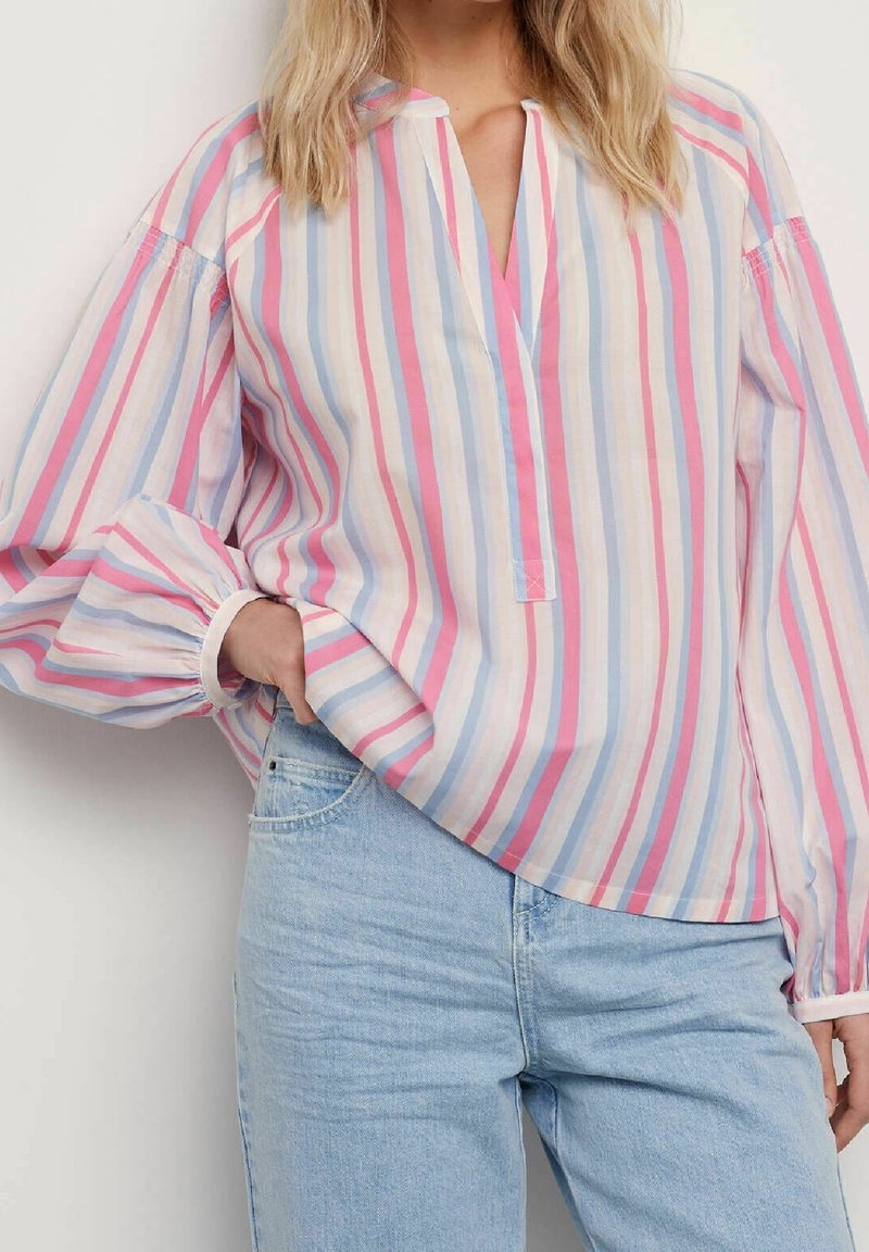Femme portant un chemisier rayé pastel à manches bouffantes, rentré dans un jean en denim bleu clair, debout avec une main dans la poche.