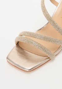 Sandale à talons en or métallique, ornée de trois fines lanières décorées de strass, à bout carré et à texture lisse et brillante.