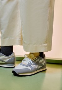 Person som bär ljusbeiga byxor och gråvita Reebok-sneakers står på grön golv.