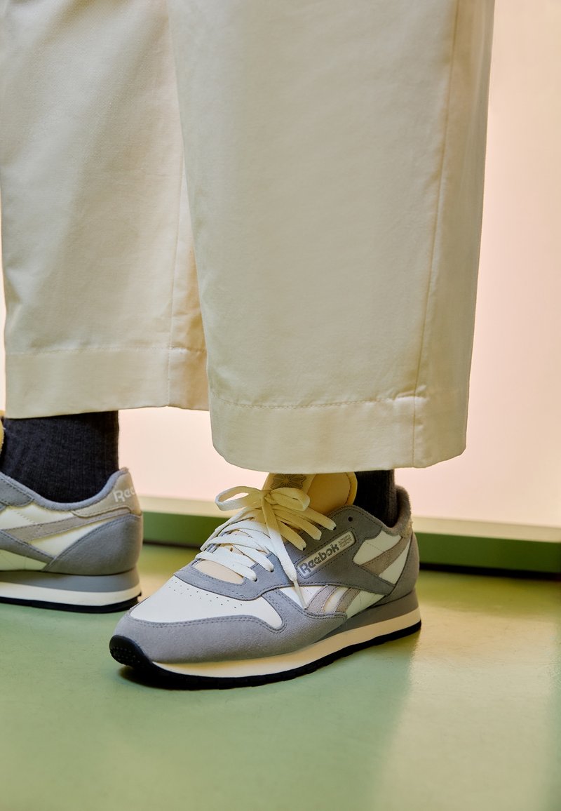 Person som bär ljusbeiga byxor och gråvita Reebok-sneakers står på grön golv.