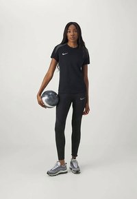 Melnā sportiskā Nike T-kreklā un legingos ar teksturētu rakstu, apģērbs papildināts ar sudraba un melnām sporta kurpēm, turēdams atbilstošu bumbu.