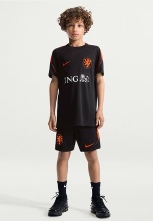 Enfant aux cheveux bouclés portant le maillot de football noir et orange des Pays-Bas, debout avec les bras détendus devant un fond clair uni.