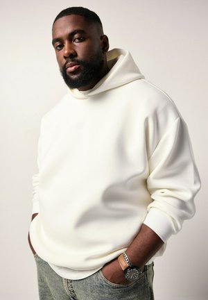 Skægget mand iført en hvid hoodie og jeans med hænderne i lommerne, bærende et ur og et armbånd på venstre håndled mod en lys baggrund.