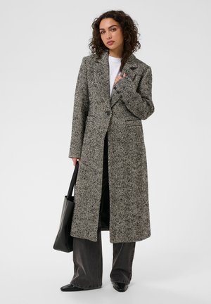 Femme portant un long manteau noir et blanc à motif chevrons, un pantalon noir à jambes larges, des chaussures noires et tenant un sac à main noir.