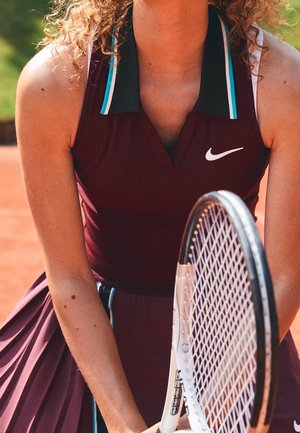 Kobieta tenisistka ubrana w bordowy strój Nike, trzymająca rakietę tenisową na korcie ziemnym, w trakcie akcji przygotowująca się do uderzenia piłki.