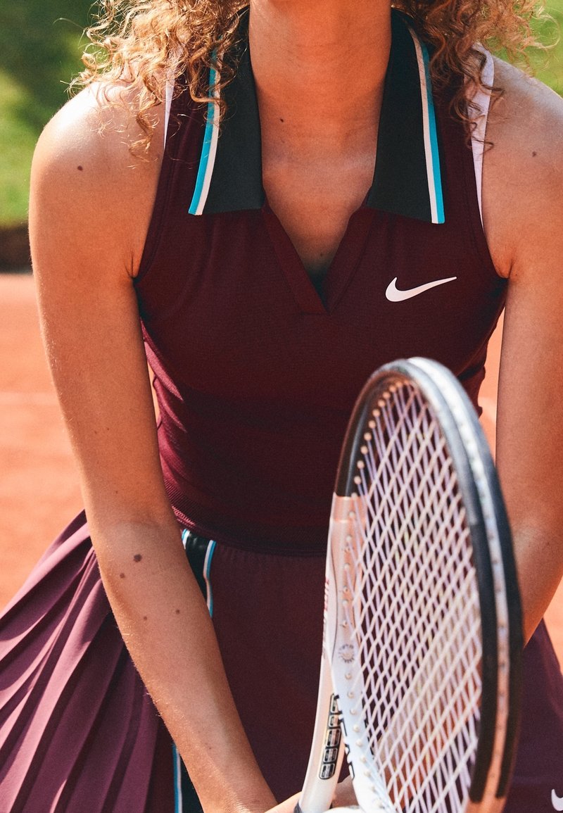 Joueuse de tennis féminine portant une tenue bordeaux Nike, tenant une raquette de tennis sur un court en terre battue, en pleine action, prête à frapper la balle.