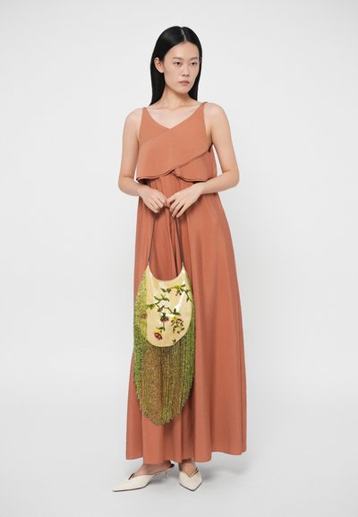 Γυναίκα σε αμάνικο μέγιστο φόρεμα σε πορτοκαλί-καφέ απόχρωση που κρατάει κίτρινη τσάντα με κορδόνια και πράσινες φούντες, καθώς και λευκές μυτερές γόβες.