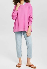 Felpa rosa con vestibilità comoda, maniche raglan e fessure laterali, indossata sopra una camicia a quadri blu e jeans cropped azzurri. Sandali marroni.