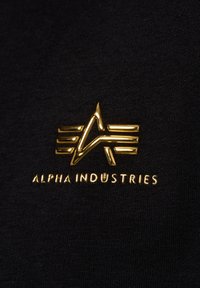 Tessuto nero con una texture liscia, caratterizzato da un emblema dorato a forma di stella e righe, con la scritta "ALPHA INDUSTRIES" in un carattere grassetto sottostante.