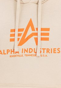 Bežna kapuca z velikim oranžnim logotipom "Alpha Industries" in oblikovalskimi elementi, z vrvicami in mehkimi, teksturiranimi materiali.
