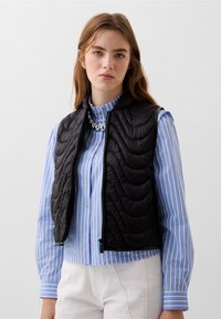 Gilet imbottito nero con design senza maniche e colletto alto, indossato sopra una camicia button-up a righe blu e bianche. Dettagli in metallo argentato.