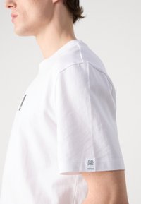 T-shirt en coton blanc à manches courtes, avec un logo sur le côté gauche à l'avant et une étiquette de marque sur la manche. Texture lisse, coupe classique.