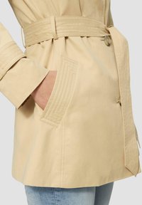 Beige trenčkot s páskem v pase, s diagonálně šitými kapsami, hladkým materiálem a knoflíkovým zapínáním na pravé straně.