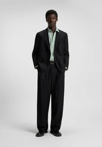Black suit with a double-breasted jacket, light green shirt, wide-leg trousers, and black shoes. Smooth fabric, minimalist design, no patterns.