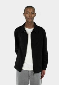 Svart semsket skinnjakke med glidelås foran, krage og brystlomme, kombinert med en hvit t-skjorte og grå jeans. Glatt tekstur, uformelt design.