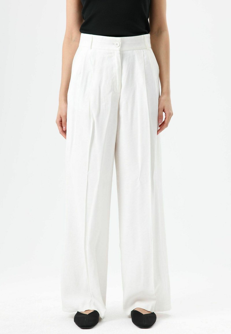 Pantalon blanc à taille haute, jambes larges, avec plis devant et fermeture à un seul bouton, en tissu léger. Chaussures plates noires portées.