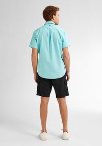 Korte mouwen, overhemd met kraag in lichtteal stof met een rugplooitje. Gecombineerd met zwarte cargoshorts en beige schoenen.