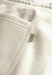 Tissu beige clair avec une texture lisse, comportant une poche latérale et une petite étiquette de marque avec un logo de couronne. Coutures visibles.