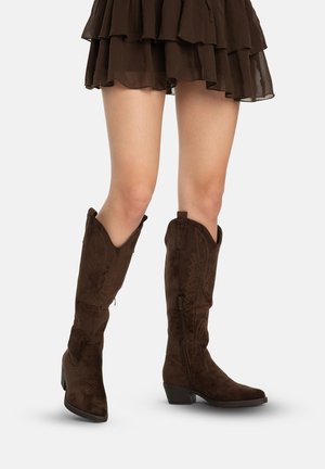 Beine in kniehohen, dunkelbraunen Wildleder-Cowboystiefeln mit Stickereien und seitlichen Reißverschlüssen, kombiniert mit einem mehrlagigen braunen Rock.