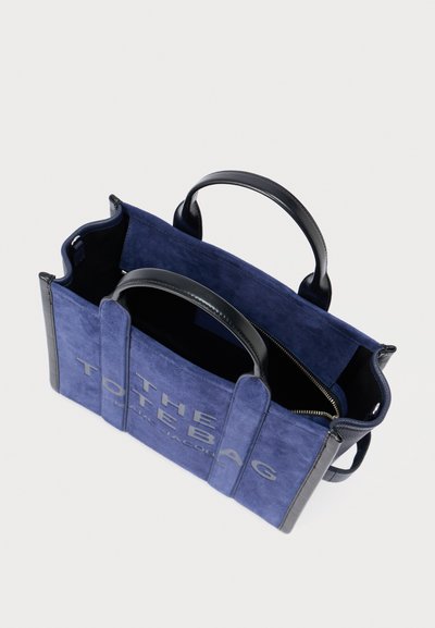 Bolso tote de ante azul marino con detalles de cuero negro, dos asas y un bolsillo con cremallera; presenta un texto en relieve y en negrita que dice "THE TOTE BAG".