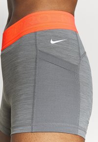 Pantaloncini sportivi grigi con finitura testurizzata, dotati di fascia elastica arancione in vita e dettaglio logo bianco. Tasca laterale integrata.