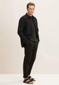 Zwarte button-up shirt over een donkere trui, gecombineerd met zwarte broeken en zwarte sandalen. Minimalistisch ontwerp met een relaxte pasvorm.