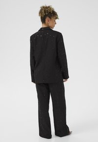Schwarzes Anzugset mit strukturiertem Design und kleinen Mustern. Der Blazer hat einen klassischen Kragen und Knopfmanschetten; die Hose hat weite Beine.