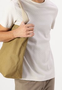 Bežové strukturované tričko s kulatým výstřihem, doplněné hořčičkovou plátěnou taškou s logem. Hnědé kalhoty dotvářejí celý outfit.